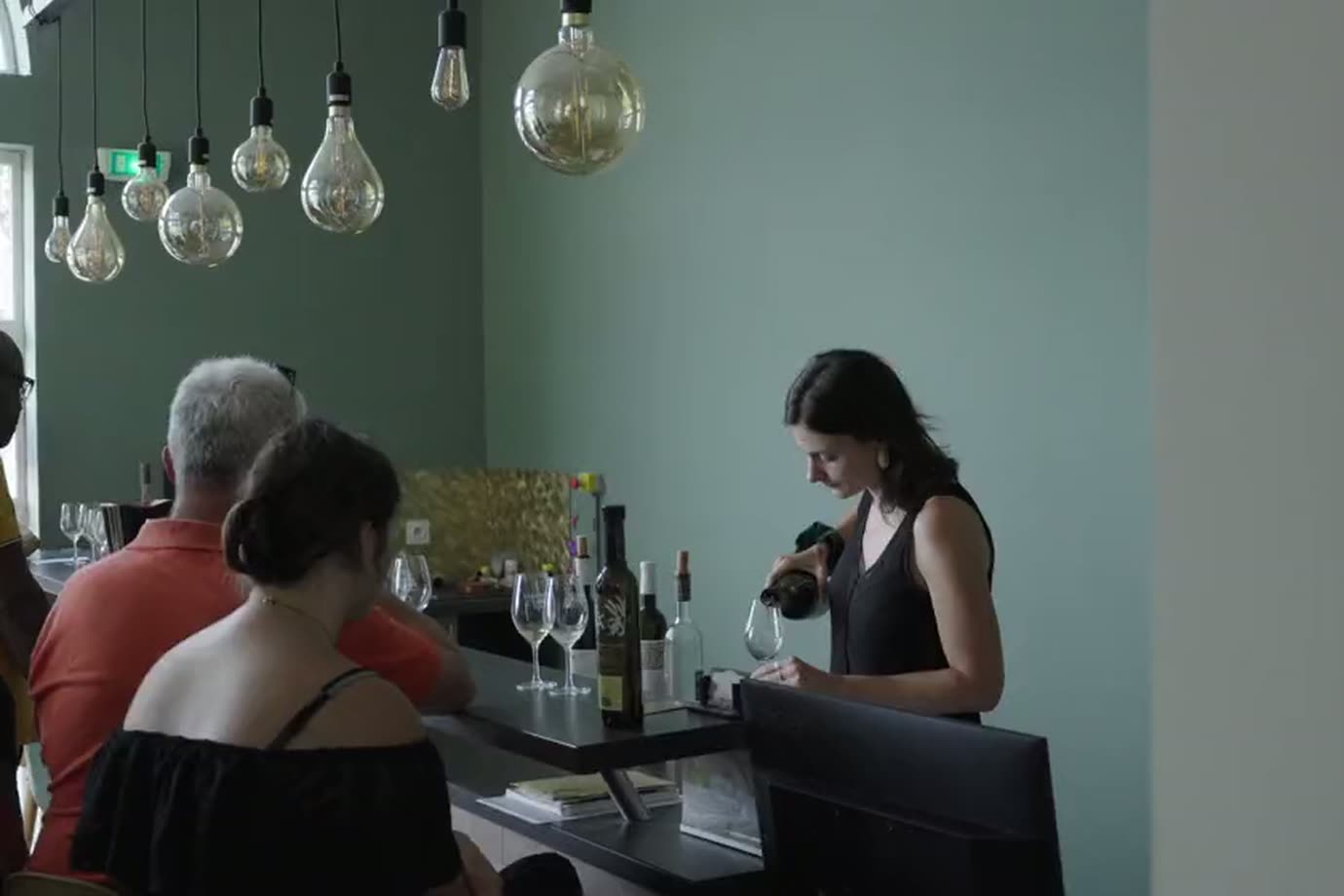 Salle de dégustation : des visiteurs au comptoir sous des suspensions.
