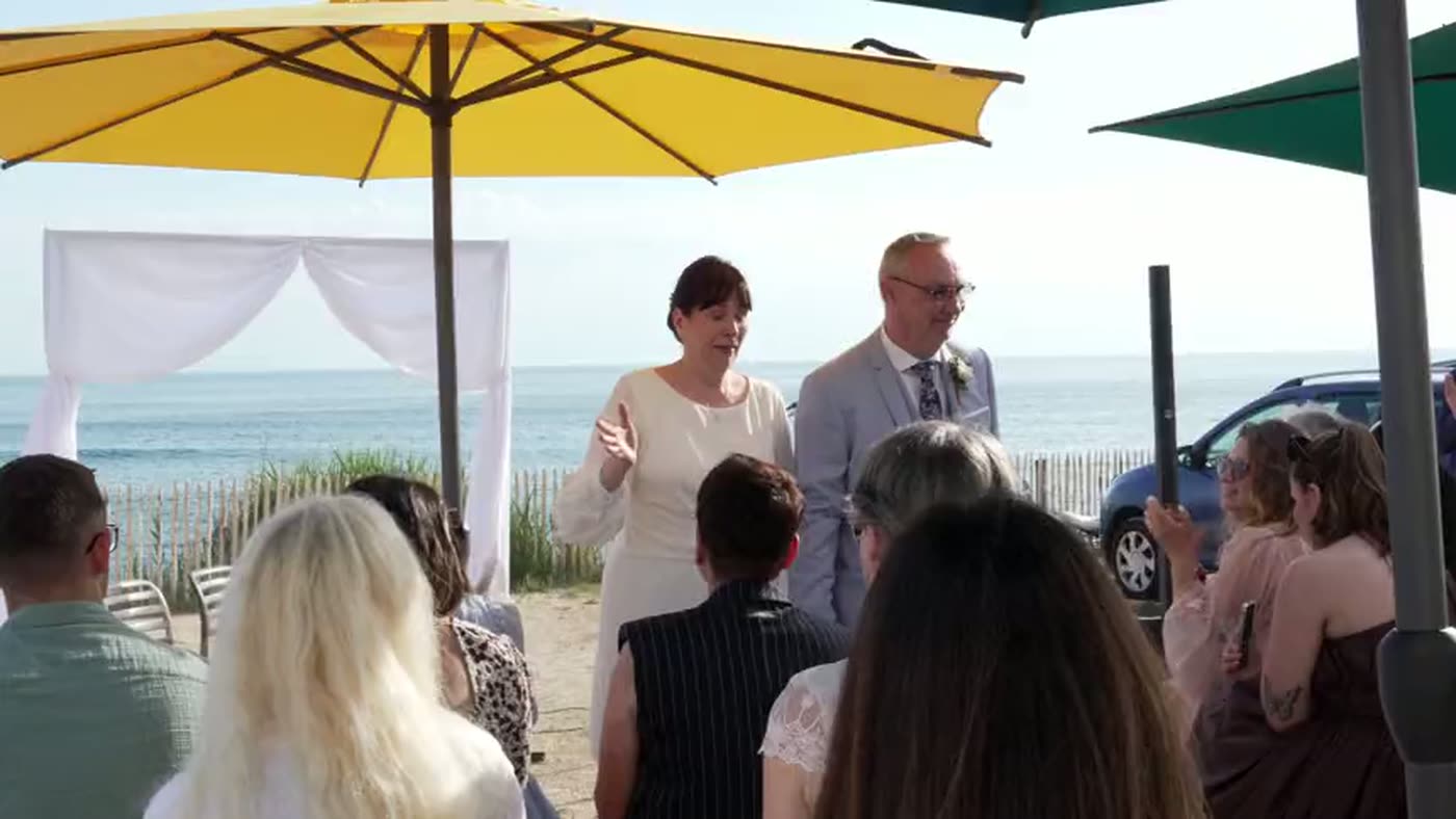 Cérémonie en bord de mer : les invités sous des parasols, la plage et l'eau derrière.